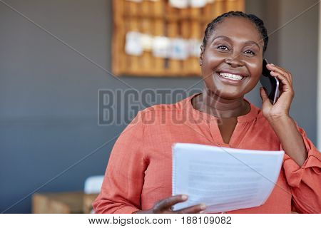 Portrait of a smiling young African businesswoman reading paperwork and talking on a cellphone while standing in large a modern office