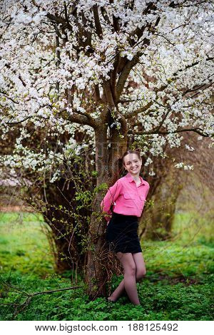 Young Beautiful Girl In A Pink Shirt Standing Under Blossoming Apple Tree And Enjoying A Sunny Day.