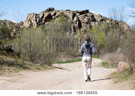 Female Hiker Is Walking To The Rocks. White Road, Bushes And Trees, Springtime