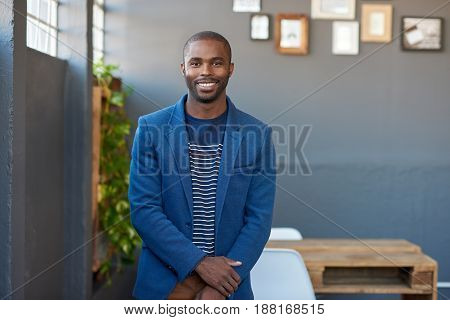 Portrait of a young African businessman in a blazer smiling confidently while standing alone in a large modern office