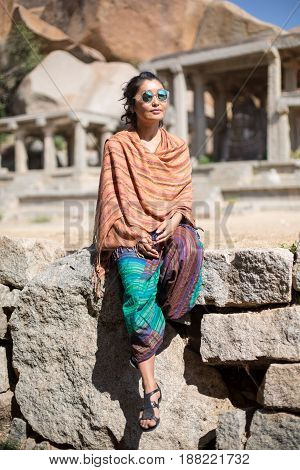 A girl in a cloak sits on a stone wall against a ruin background