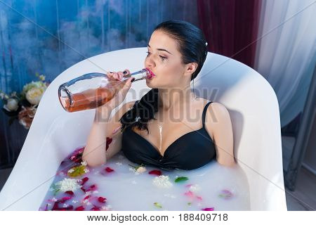 Sexy brunette drunk girl in a hot bath wearing black sexual lingerie. She is drinking champagne straight from the bottle alone. break up in a relationship concept.