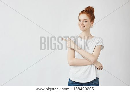 Beautiful ginger girl smiling pointing finger in side looking at camera. Copy space. Isolatedon white background.
