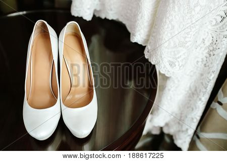 Elegant stylish white wedding shoes on wooden table with a dress in background. Morning preparetion.
