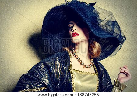Portrait of a gorgeous young woman in elegant broad-brimmed hat and luxurious dress over golden background. Vintage style. Beauty, fashion.