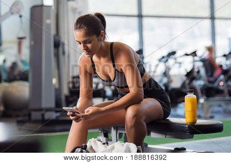 Young woman athlete using cell phone at gym. Latin woman in sportswear checking phone while resting after workout on bench. Beautiful fit girl messaging with smartphone at fitness centre.