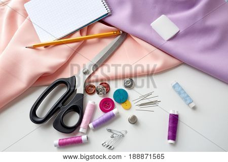 needlework and tailoring concept - scissors, sewing buttons, spools of thread, cloth and notepad with pencil