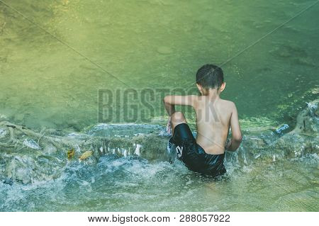 Little Boy Crossing A River Full Of Stones Carefully And Teetering In Waterfall At Erawan National P