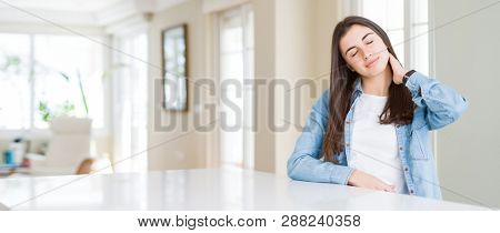 Wide angle picture of beautiful young woman sitting on white table at home Suffering of neck ache injury, touching neck with hand, muscular pain