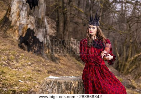 Art Photography. Mysterious Medieval Queen In Red Dress And Spiky Black Crown Posing With Ancient Bo