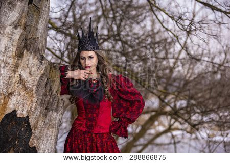 Art Photography. Beautiful Fairy Princess In Red Dress And Spiky Black Crown In Forest In Early Spri