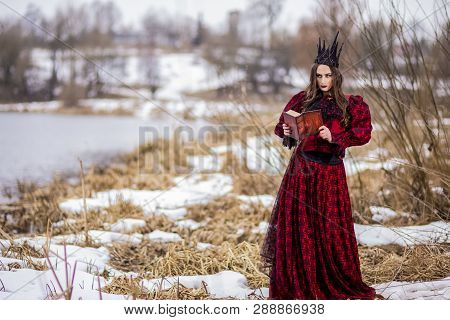 Mysterious Fairy Princess In Red Dress And Black Crown Reading Old Book. Posing In Forest Outdoors.h