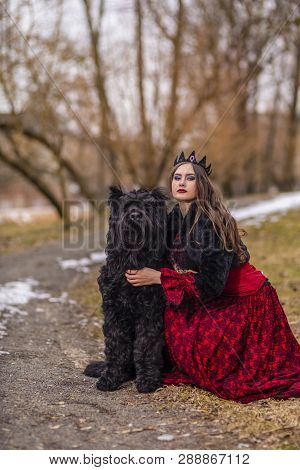 Beautiful Princess In Red Dress And Black Fur Jacket Posing In Crown Along With Her Dog In Forest Du