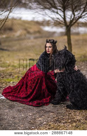 Beautiful Princess In Red Dress And Black Fur With Crown And Her Dog In Forest During Early Spring. 