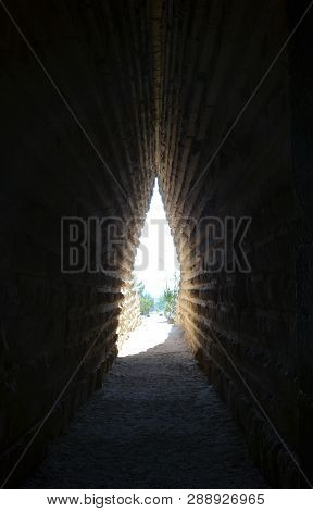 Path Through A Dark Narrow Tunnel In Old Stone Ruins. Sunlight And Green Trees At The End Of The Tun