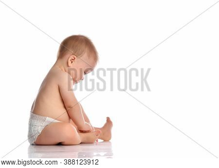 Cute Little Baby In Diapers Examines The Feet And Toes With Interest While Sitting On The Floor. Stu