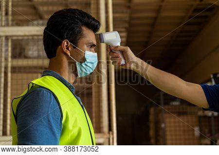 Young Indian Factory Warehouse Worker Wearing Face Mask Getting Body Temperature Check With Infrared
