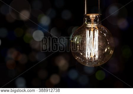 Glowing Vintage Old One Tungsten Lightbulb Hanged On Ceiling Against Black Wall Bokeh Background. Re