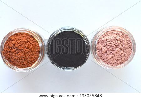 Three different mineral clays for facial masks - pink clay, red clay and activated charcoal in glass jars top view on white