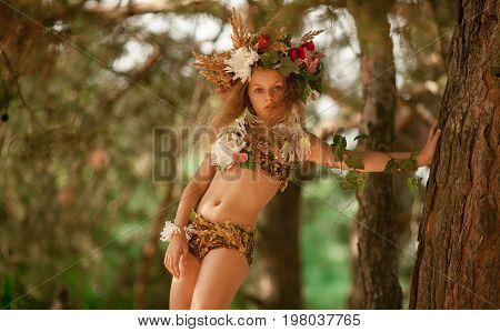 Beautiful little girl in image of nymph dryad with floral head wreath stands near tree in magical fairytale forest .