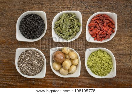 collection of superfoods in small ceramic bowls against rustic wood: black cumin seeds, macadamia nuts, hemp seed hearts, goji berry, stevia herb and chia seeds