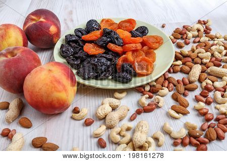 On a light table a plate of dried apricots and prunes. Near it are ripe peaches and а scattered nuts. A mixture of nuts consists of peeled and peanuts almonds and cashews.