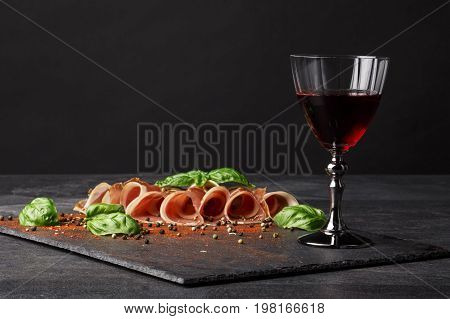 A wineglass filled red wine, salted and raw balyk, spicy basil on a black background. Prosciutto and semi sweet wine. A glass of wine and a pile of cut ham on a black background. Copy space.