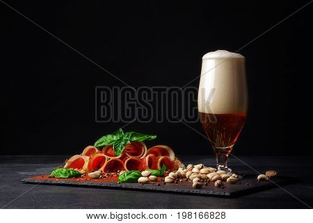 A tall and huge glass filled with golden cold beer and salted pistachios, cut twisted pieces of prosciutto on a black background. Copy space. Pistachios and a plate of sliced balyk pieces.