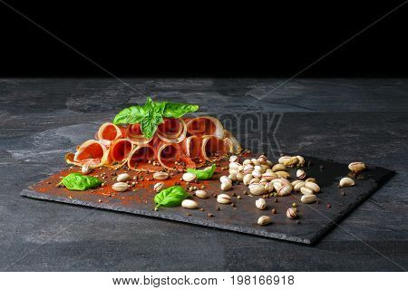 A plate of perfectly sliced prosciutto with green leaves of basil and pepper on a black background. Cured balyk with pistachios. Sliced smoked bacon with pepper. Raw and salt meat. Copy space.