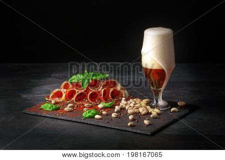 A composition of a glass of dark beer, pistachios and a plate of perfectly sliced balyk with spinach on a dark background. Cured prosciutto with a basil and a goblet of alcoholic beer with foam.