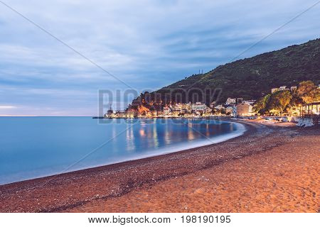 Petrovac village night scene with montenegrin houses, Castello fortress, seaview and evening lights reflected in the water of Adriatic sea. Touristic town Petrovac harbor panorama, Montenegro.