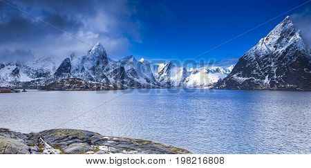 Travel Concepts. One Small Village on Seashore Coastline in Norway Against Mountains Covered With Snow.Horizontal Image Orientation