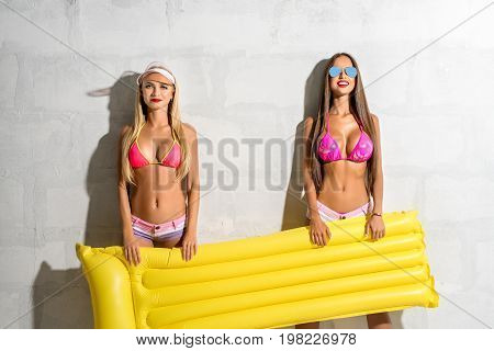 Two sexy girls wearing sunglasses, bicinis posing near sunny brick white wall holding yellow swimming air mattress
