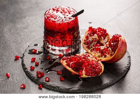 A close-up of a colorful alcohol drink with garnet juice, ice and black straw. A raw and tasteful pomegranate and a glass of red juice on a light gray background. Delicious garnet products on a tray.