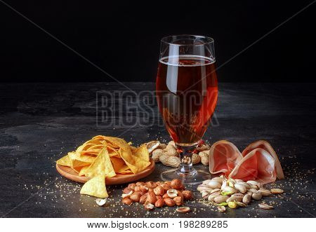 A beautiful composition of different snacks and a pint of saturated brown beer on a black table background. Peanuts, pistachios and a plate of chips. A beer with a perfectly sliced balyk. Copy space.