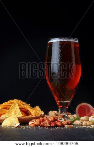 A close-up picture of a tall flass full of golden cold beer on a table. A beer with spicy snacks: salty pistachios, closed and cracked peanuts, yellow nachos chips and cut ham on a black background.
