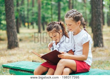 Two schoolgirls of junior classes sit on a bench and read a book. Back to school