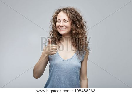 Portrait of a happy young Caucasian successful woman giving in grey casual shirt showing thumb up or like gesture looking and smiling camera with happy expression. Human emotions face expressions.