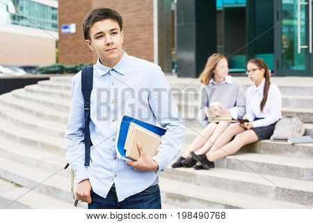 Portrait of teenage boy holding books standing outside school building, looking away pensively