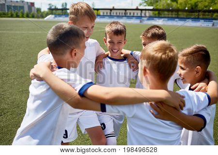 Team of boys in uniform embracing smiling happily during practice on football field