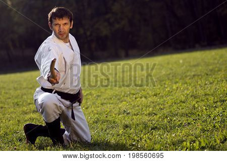 The man in white kimono and black belt is practicing martial arts with wooden sword outdoors on the grass.
