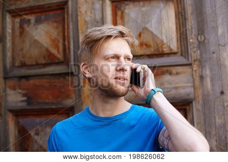 Picture of seious young blonde man with beard talking on generic mobile phone standing outdoors at wooden door calling to taxi service to get a cab. Attractive guy having phone conversation