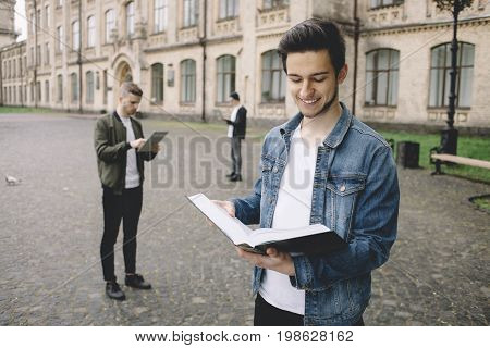 These young men are standing on the road near the university. One of them are reading the book and smiling, another one is concentrated on something while looking on his tablet. The last one is searching for some information on his laptop. Close up