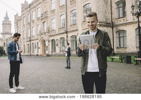 These young men are standing on the road near the university. One of them are reading the book and smiling, another one is concentrated on something while looking on his tablet. The last one is searching for some information on his laptop. Close up