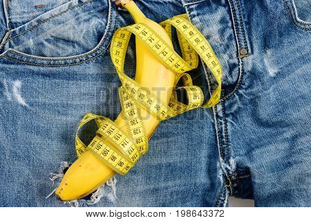 Health and male sexuality concept. Banana wrapped with yellow measure tape on jeans selective focus. Mens denim pants crotch with banana imitating male genitals. Jeans zipper and pocket close up.