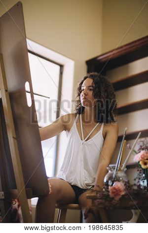 Smiling artist girl with black curly hair in white shirt and jeans shorts painting an intresting picture on canvas in her artstudio during the daylight