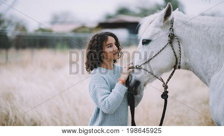 Attractive and beautiful woman with black culy hair in a light blue coat tenderly stroking a gorgeous white horse. They are walking outside. Close up
