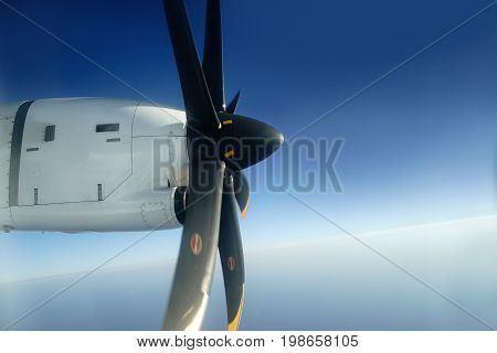 Outside view from window seat of small airplane propeller engine attached to the wings with its black blades rotating high up in the clear blue sky.