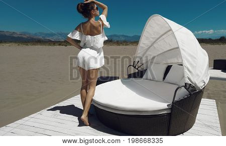 Fashionable glamour woman Luxury travel vacation. Fashion tanned brunette in white by beach sofa on sand before blue sky. Girl enjoying the sun carefree happy.