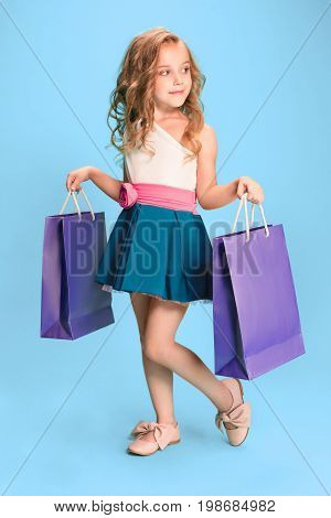 The cute little caucasian brunette girl in hat holding shopping bags on blue studio background